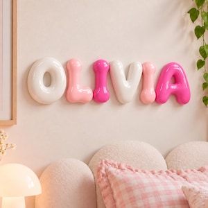 May include: A wall-mounted decorative sign spelling the name OLIVIA in three-dimensional, glossy, balloon-style letters. The letters alternate in colour between white, light pink, and bright hot pink, creating a smooth, rounded appearance.