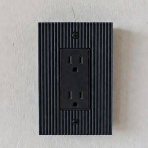 May include: A black electrical socket with a textured, vertically-lined faceplate. The socket has two standard three-pin receptacles and is secured to the wall with two screws. The faceplate has a rectangular shape.
