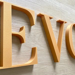 May include: Three-dimensional capital letters spelling the word EVOLVE in a metallic gold finish. Each letter features a clean, serif-style typeface with a smooth, matte texture and a consistent depth, casting subtle shadows on the surface below.