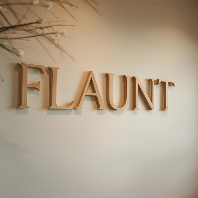 May include: A set of three-dimensional, light beige wooden letters spelling the word FLAUNT, mounted horizontally on a plain, light-colored wall. The letters feature a clean, serif typeface with a smooth finish and cast soft shadows below.