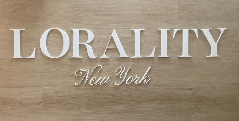 May include: White three-dimensional lettering spelling LORALITY in a bold serif font, positioned above the words New York in a white cursive script. The letters are mounted on a light brown wood-grain surface with a horizontal plank pattern.