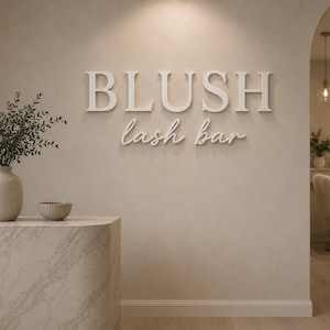 May include: A beige wall featuring the white 3D text BLUSH lash bar. To the left, a marble-textured counter holds a beige vase with green foliage and a small bowl. An arched doorway reveals a salon chair and a circular mirror with backlighting.