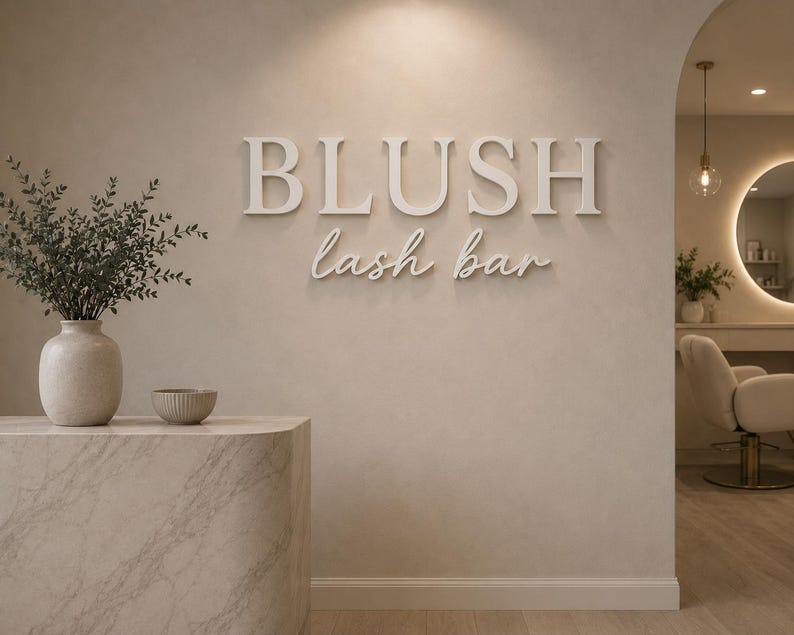 May include: A beige wall featuring the white 3D text BLUSH lash bar. To the left, a marble-textured counter holds a beige vase with green foliage and a small bowl. An arched doorway reveals a salon chair and a circular mirror with backlighting.