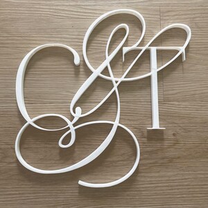 May include: A white wall-mounted sign featuring the letters C and T connected by an ampersand symbol. The letters are rendered in a flowing, cursive script font with thin, elegant lines, creating a continuous decorative monogram design.