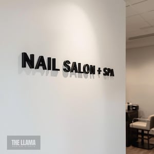 May include: A wall-mounted sign featuring three-dimensional black lettering that reads NAIL SALON + SPA. The letters are mounted on a plain white wall, creating a distinct shadow effect beneath each character due to the overhead lighting.