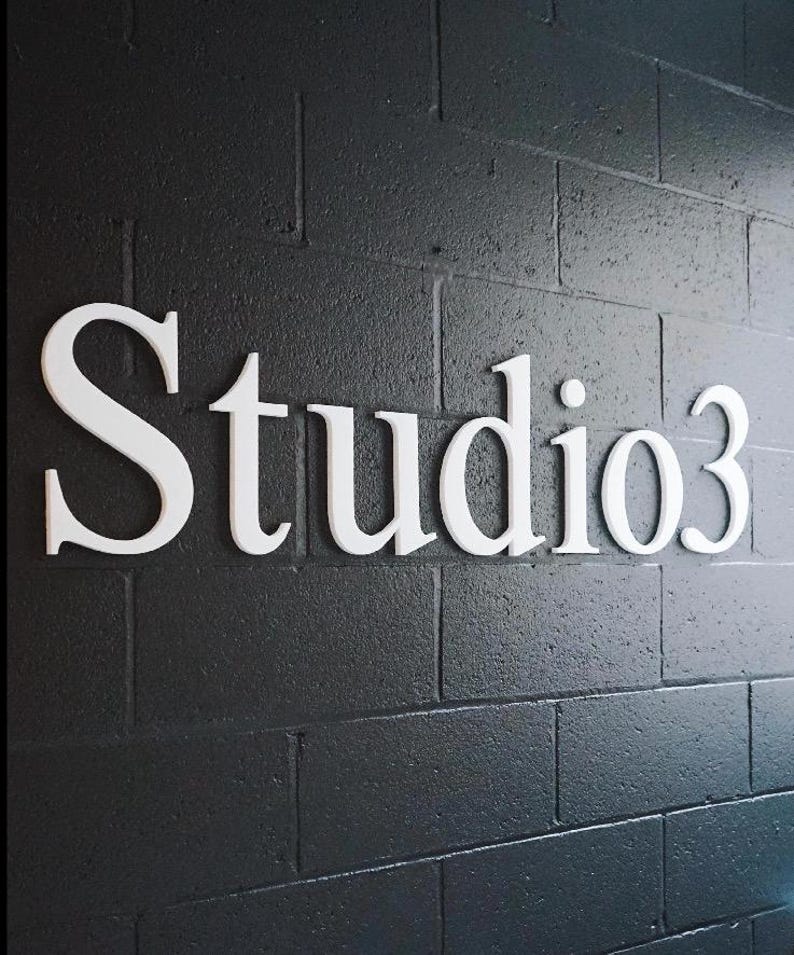 May include: A three-dimensional sign featuring the white text Studio3 mounted on a dark gray, textured cinder block wall. The letters are capitalized and lowercase, displaying a serif font style with clean edges and a matte finish.