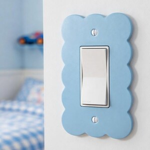 May include: A pale blue, cloud-shaped light switch cover with a white switch. The cover is fixed to a white wall. The room has a blue and white theme, with a bed and toy car visible in the background.