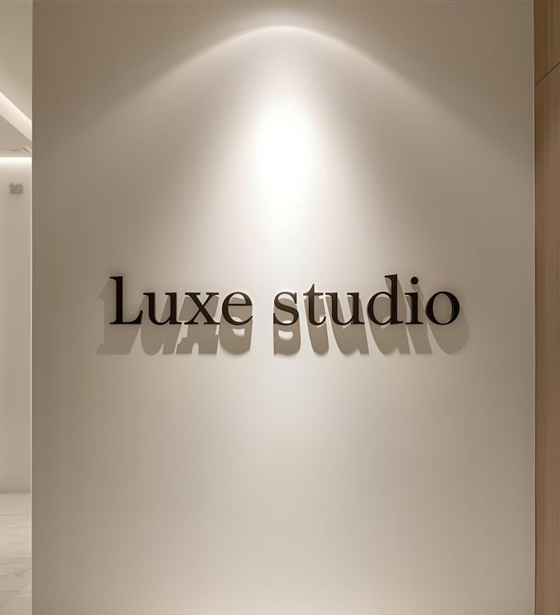 May include: A wall-mounted sign featuring the words Luxe studio in dark, three-dimensional lettering. The letters are mounted on a smooth, off-white wall surface, creating distinct shadows beneath each character under overhead lighting.