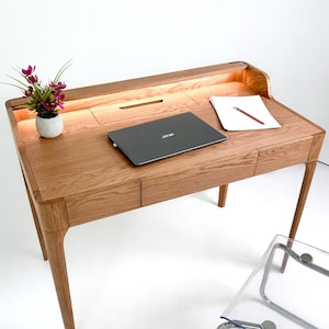 May include: A wooden desk with a built-in light, two drawers, and a laptop. A small potted plant with purple flowers sits on the desk. A sheet of paper and a pencil are also on the desk. The desk has a modern design with tapered legs.