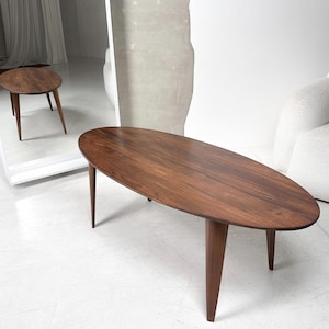 Pode incluir: Uma mesa de centro oval de madeira escura com uma superfície lisa e três pernas. A mesa é feita de madeira maciça e tem um acabamento natural.