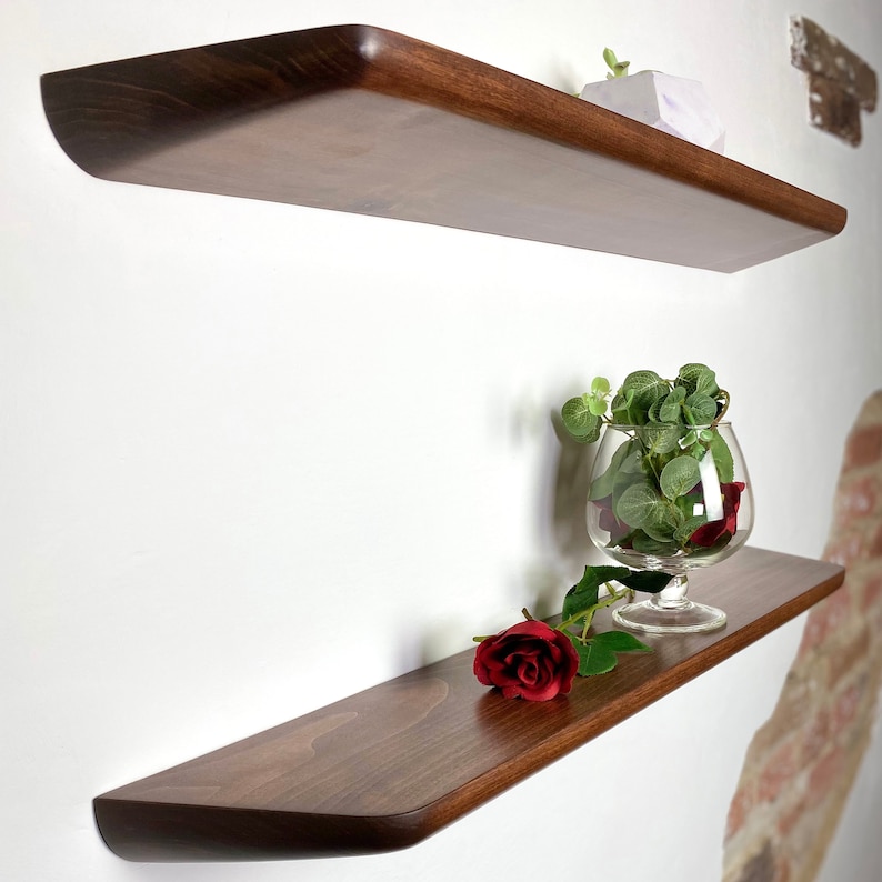 May include: Two dark brown wooden floating shelves mounted on a wall. The bottom shelf holds a clear glass goblet filled with green leaves and a single red rose lying flat on the surface. The top shelf features a small white geometric object.