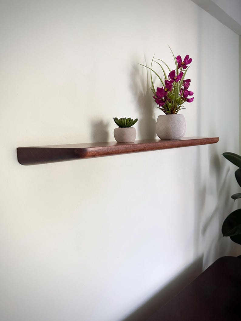 May include: A long, dark brown wooden floating shelf mounted on a wall. On the shelf sits a small, light gray pot containing a green succulent and a larger, light gray rounded pot holding a bouquet of vibrant magenta flowers with green leaves.
