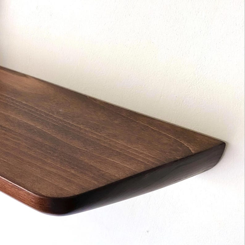 May include: A wall-mounted floating shelf crafted from dark brown wood with a visible natural grain pattern. The shelf features a smooth, rounded front edge and a rectangular shape, extending horizontally from a plain white wall surface.