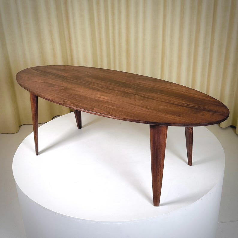 May include: Oval-shaped wooden coffee table with a dark brown finish. The table has four tapered legs. The table is displayed on a white pedestal against a neutral background.