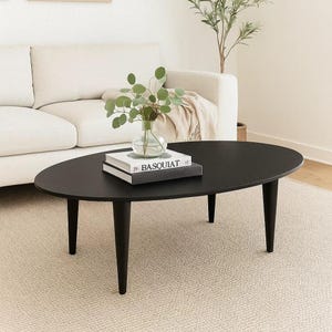 May include: A black oval coffee table with four tapered legs. A clear glass vase with green foliage and two books are on the table. The table is on a beige rug in front of a white sofa.