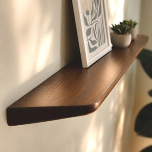 May include: A dark brown wooden floating shelf with a smooth, tapered edge. The shelf holds a white-framed print featuring a floral illustration in shades of beige and dark gray, alongside two small, round, light gray pots containing green succulents.