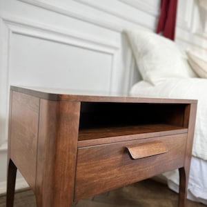 May include: A dark brown wooden bedside table featuring a smooth rectangular top, an open storage shelf located directly beneath the top surface, and a single pull-out drawer equipped with a horizontal wooden handle, supported by four curved legs.
