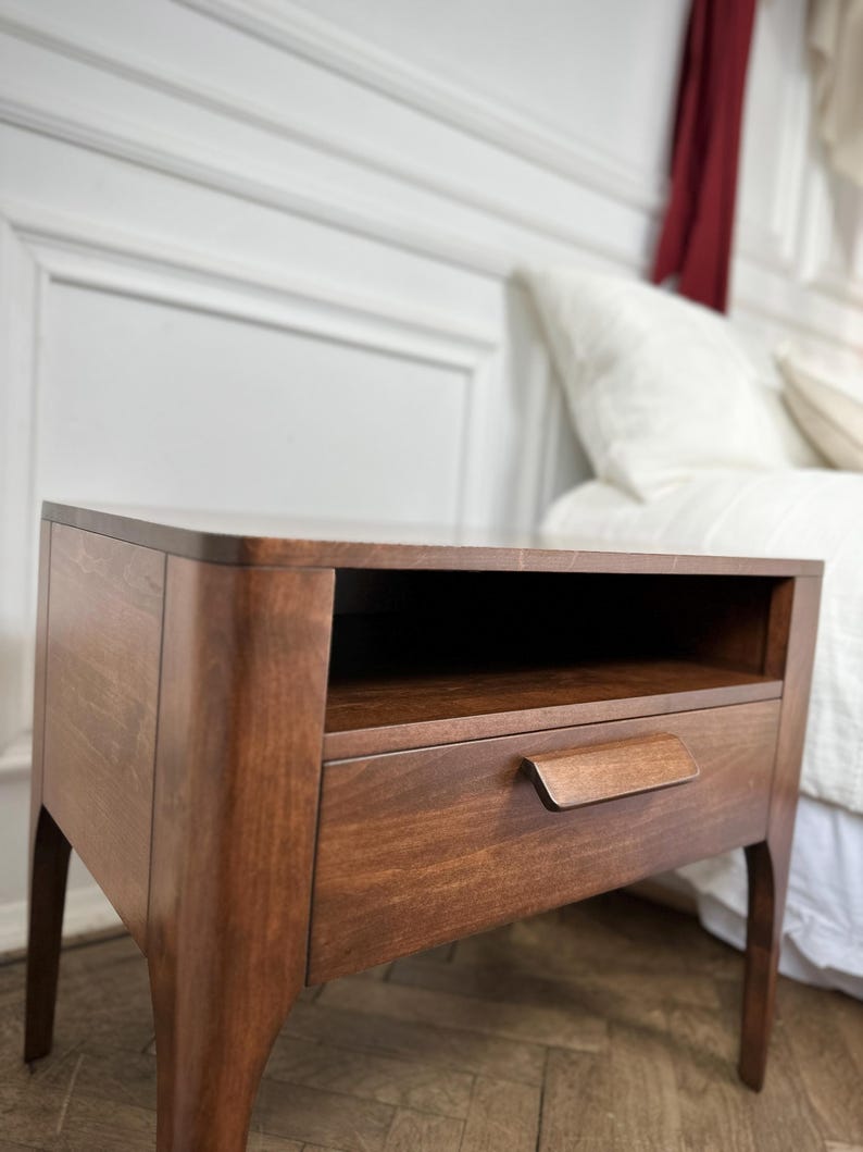 May include: A dark brown wooden bedside table featuring a smooth rectangular top, an open storage shelf located directly beneath the top surface, and a single pull-out drawer equipped with a horizontal wooden handle, supported by four curved legs.