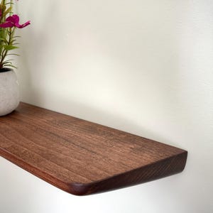 May include: A dark brown wooden floating shelf with a smooth, rounded front edge. A small, light gray ceramic pot containing artificial pink cosmos flowers sits on the left side of the shelf, which is mounted against a plain white wall.