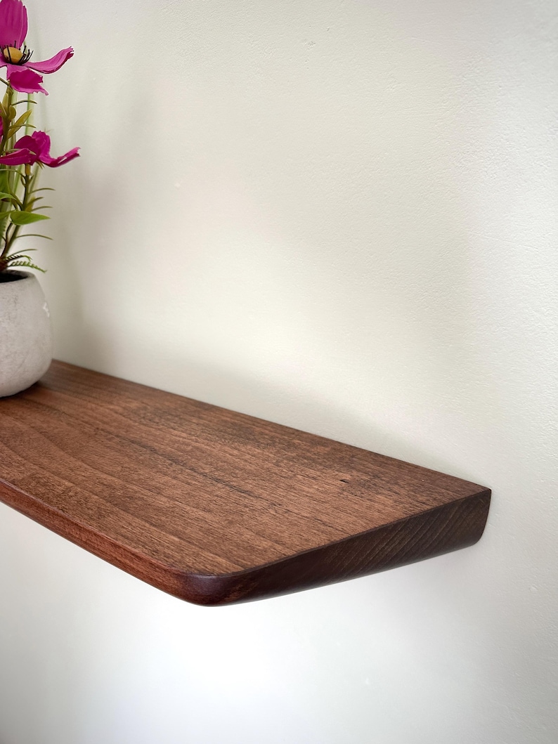 May include: A dark brown wooden floating shelf with a smooth, rounded front edge. A small, light gray ceramic pot containing artificial pink cosmos flowers sits on the left side of the shelf, which is mounted against a plain white wall.