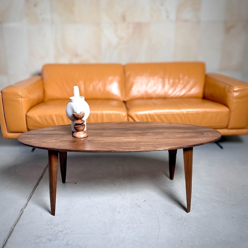 May include: Oval wooden coffee table with tapered legs. A white ceramic vase with a wooden base sits on the table. A tan leather sofa is in the background. The table is approximately 48 inches long.