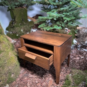 May include: A brown wooden bedside table featuring a rectangular top surface, an open storage shelf directly beneath the top, and a single pull-out drawer with a horizontal wooden handle. The piece is supported by four tapered legs.