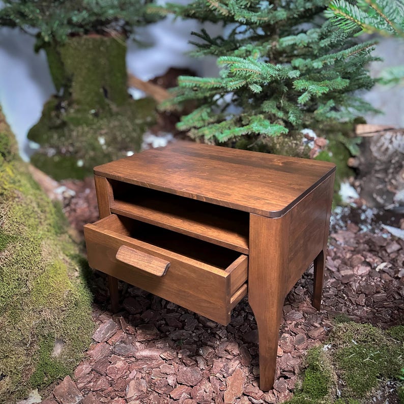 May include: A brown wooden bedside table featuring a rectangular top surface, an open storage shelf directly beneath the top, and a single pull-out drawer with a horizontal wooden handle. The piece is supported by four tapered legs.