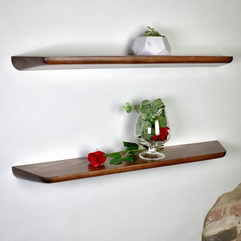 May include: Two horizontal floating shelves made of dark brown wood with a smooth finish. The top shelf holds a white geometric planter with a small green succulent. The bottom shelf features a clear glass goblet containing green leaves and a red rose.