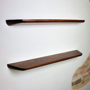 May include: Two horizontal floating shelves made of dark brown wood with a smooth finish and rounded front edges. The shelves are mounted parallel to each other on a plain white wall, showcasing a clean, linear design with visible wood grain.
