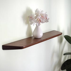 K&ouml;nnte beinhalten: Ein dunkelbraunes Holz-Wandregal mit einer glatten, abgerundeten Vorderkante. Auf dem Regal steht eine kleine, mattwei&szlig;e Keramikvase mit einem Strau&szlig; wei&szlig;er Orchideen, die zartrosa Zentren aufweisen und ein schlichtes Design zeigen.