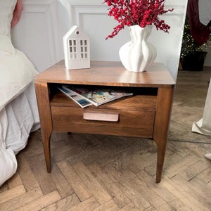 May include: A mid-century modern wooden bedside table with a single drawer featuring a horizontal handle and an open storage shelf. On top sits a white ceramic house-shaped lantern and a white fluted vase filled with bright red berry branches.