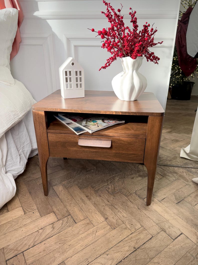 May include: A mid-century modern wooden bedside table with a single drawer featuring a horizontal handle and an open storage shelf. On top sits a white ceramic house-shaped lantern and a white fluted vase filled with bright red berry branches.
