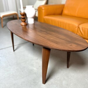 May include: A mid-century modern oval coffee table with a rich, dark wood finish. The table has a smooth surface and tapered legs. A white vase and a wooden candle holder with a white candle are on the table.