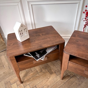 May include: Two dark brown wooden bedside tables with rounded corners, each featuring a single drawer and an open storage shelf. A white ceramic house-shaped ornament sits on the left table, which also holds a magazine titled Escape Mode.