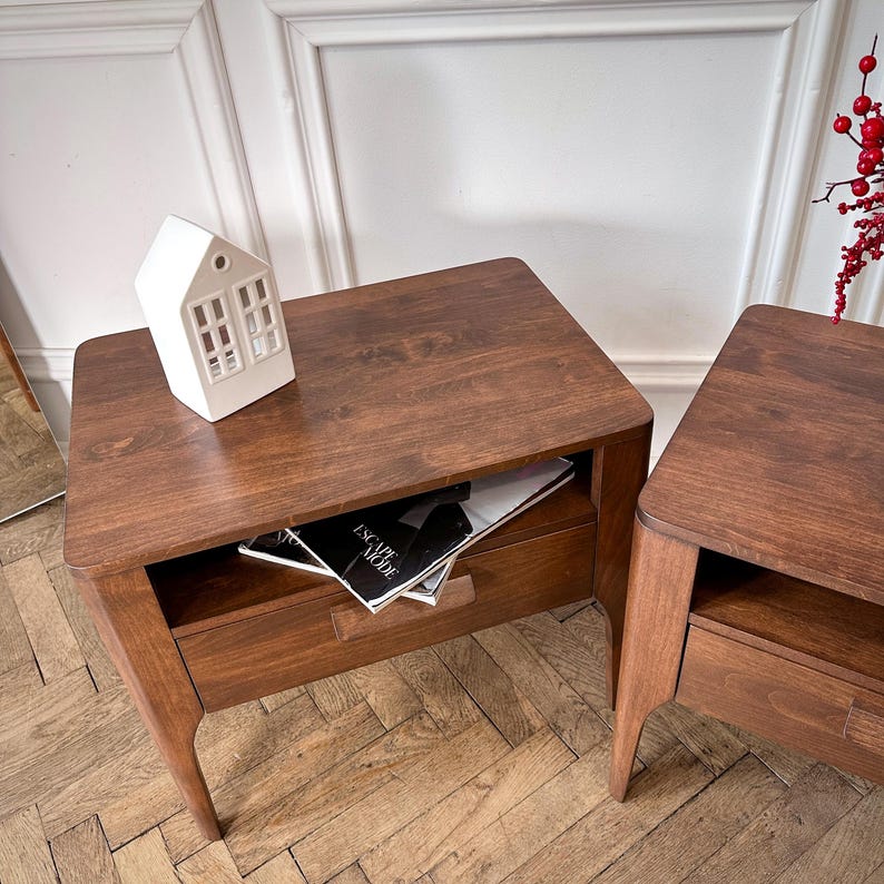 May include: Two dark brown wooden bedside tables with rounded corners, each featuring a single drawer and an open storage shelf. A white ceramic house-shaped ornament sits on the left table, which also holds a magazine titled Escape Mode.