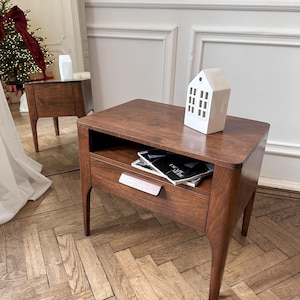 May include: A dark brown wooden nightstand with four tapered legs, an open storage shelf, and a single drawer featuring a horizontal handle. A white ceramic house-shaped decorative object sits on the top surface, and a magazine is inside the shelf.