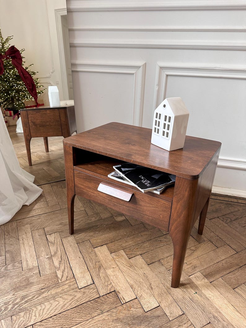 May include: A dark brown wooden nightstand with four tapered legs, an open storage shelf, and a single drawer featuring a horizontal handle. A white ceramic house-shaped decorative object sits on the top surface, and a magazine is inside the shelf.