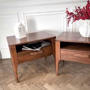 May include: Two matching dark brown wooden bedside tables with a single drawer and an open storage shelf. Each table features tapered legs and a horizontal wooden drawer pull. A white ceramic house figurine and a white vase with red berries sit on top.