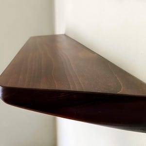K&ouml;nnte beinhalten: Ein wandmontiertes schwebendes Regal aus dunkelbraun gebeiztem Holz mit einer glatten, polierten Oberfl&auml;che. Das Regal verf&uuml;gt &uuml;ber eine rechteckige Oberseite mit abgerundeten Ecken und eine abgeschr&auml;gte Unterseite zur Wandmontage.