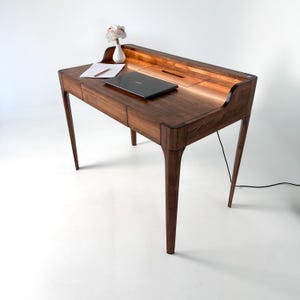 Könnte beinhalten: Ein Holzschreibtisch mit warmbraunem Finish. Der Schreibtisch hat ein geschwungenes Design mit zwei Schubladen, einem eingebauten Fach und schlanken Beinen. Ein Laptop, ein Notizblock, ein Bleistift und eine kleine Vase mit Blumen stehen auf dem Schreibtisch. Der Schreibtisch ist ca. 122 cm breit.