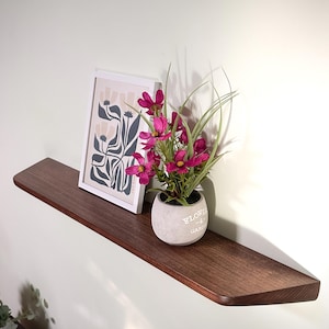 May include: A dark brown wooden floating shelf mounted on a wall. On the shelf sits a white framed print featuring a stylized floral illustration and a small grey concrete pot containing vibrant magenta artificial flowers and green grass blades.