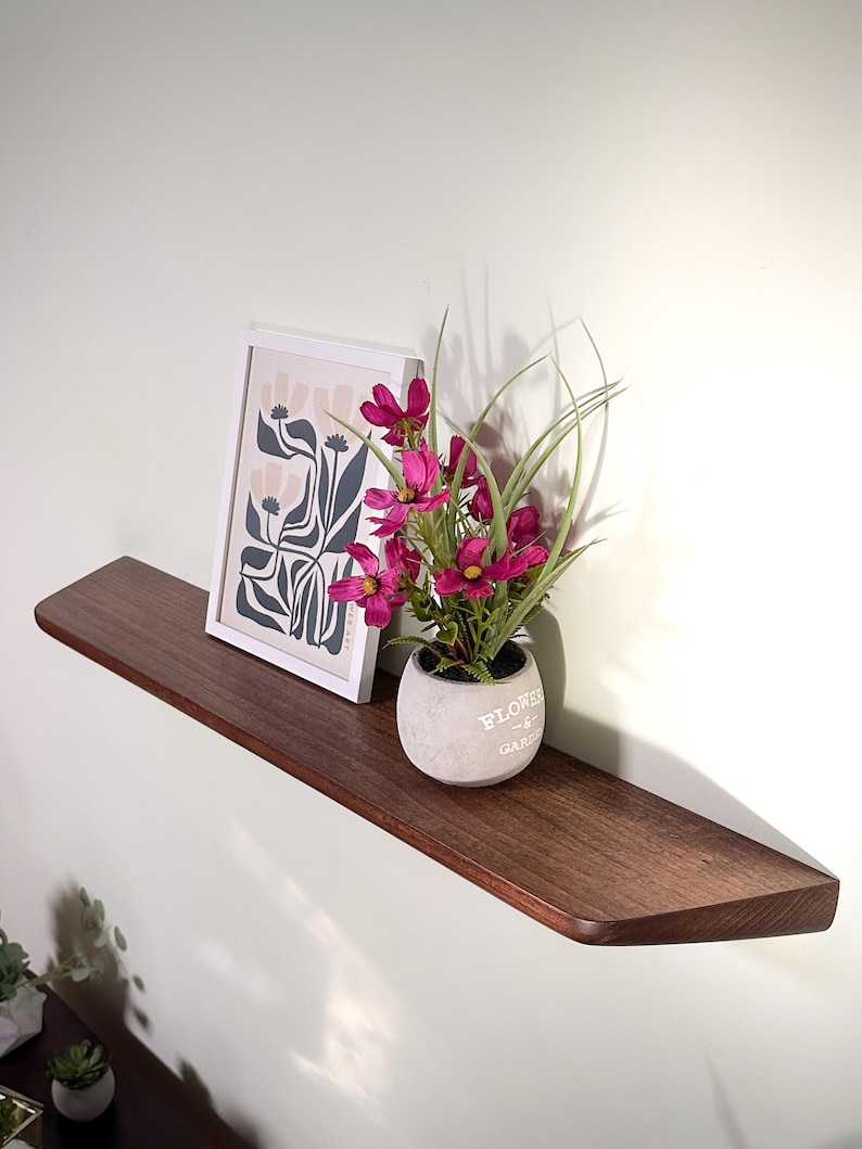 May include: A dark brown wooden floating shelf mounted on a wall. On the shelf sits a white framed print featuring a stylized floral illustration and a small grey concrete pot containing vibrant magenta artificial flowers and green grass blades.