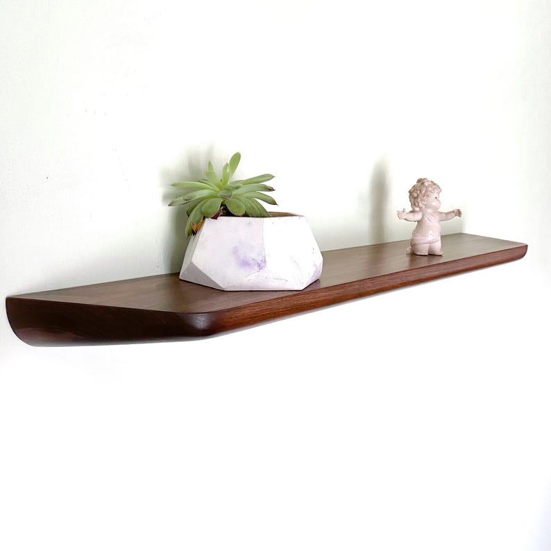 May include: A long, dark brown wooden floating shelf with rounded edges. On the shelf sits a white, geometric-shaped planter containing a green succulent plant, and a small, light-colored figurine of a person with arms outstretched.