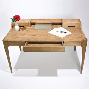 Peut inclure: Un bureau en chêne clair avec une étagère intégrée, un tiroir et un petit compartiment. Un ordinateur portable, des papiers et des ustensiles d'écriture sont sur le bureau. Un petit vase blanc avec une rose rouge est posé sur le bureau.