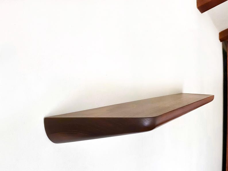 K&ouml;nnte beinhalten: Ein wandmontiertes schwebendes Regal aus dunkelbraunem Holz mit einer glatten, polierten Oberfl&auml;che. Das Regal verf&uuml;gt &uuml;ber eine rechteckige Oberseite mit abgerundeten Ecken vorne und einen dicken, festen Sockel, der zur Wand hin leicht zul&auml;uft.