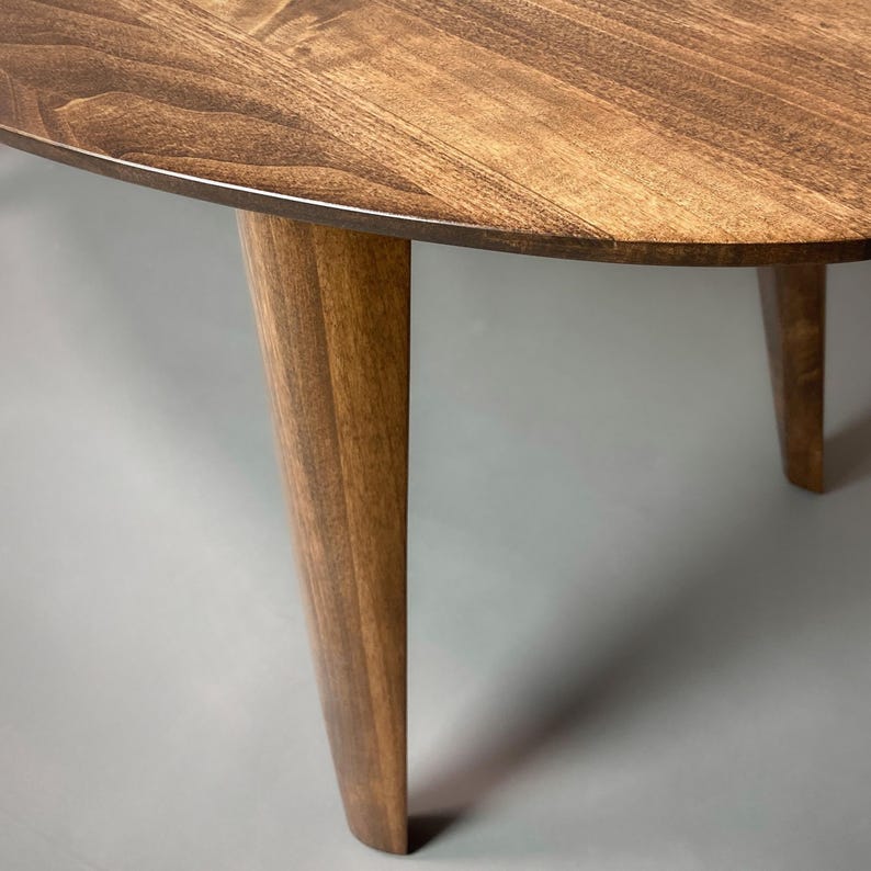 May include: A wooden oval coffee table with a dark brown finish. The table has four tapered legs. The tabletop has a visible wood grain pattern. The table is set against a gray background.