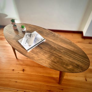 May include: Oval-shaped wooden coffee table with a dark brown finish. The table has three angled legs and is adorned with a small succulent plant and a stack of magazines. The magazines have the word "TIMELESS" printed on them.