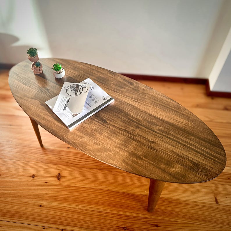 May include: Oval-shaped wooden coffee table with a dark brown finish. The table has three angled legs and is adorned with a small succulent plant and a stack of magazines. The magazines have the word "TIMELESS" printed on them.