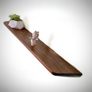 May include: A long, rectangular wooden shelf with a smooth, dark brown finish and rounded edges. A small, light-colored succulent in a geometric, light-purple pot sits on the left side, and a small, light-colored figurine stands nearby.