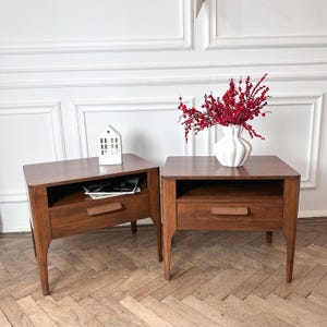 May include: Two matching dark brown wooden bedside tables, each featuring a single drawer with a horizontal handle and an open storage shelf above. A white ceramic vase filled with red berries sits on one, and a small white house model on the other.